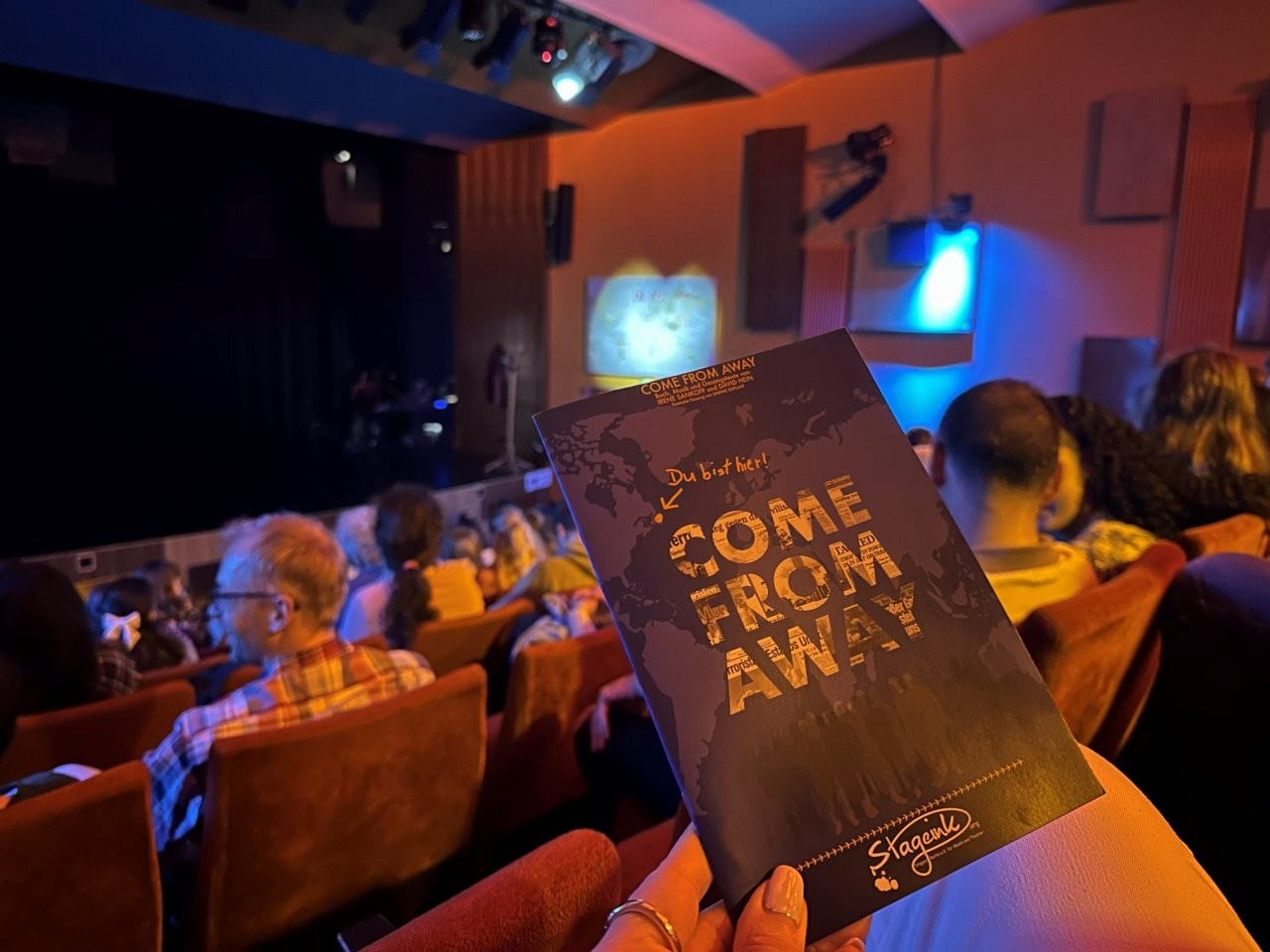 Ein Programmheft des Musicals „Come From Away“ wird im Zuschauerraum eines Theaters gehalten, während Besucher auf ihren Plätzen sitzen.